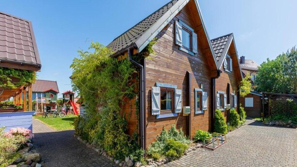 Une maison en bois avec un toit en pente, une allée pavée et un jardin agrémenté d’arbustes et d’un petit arbre.