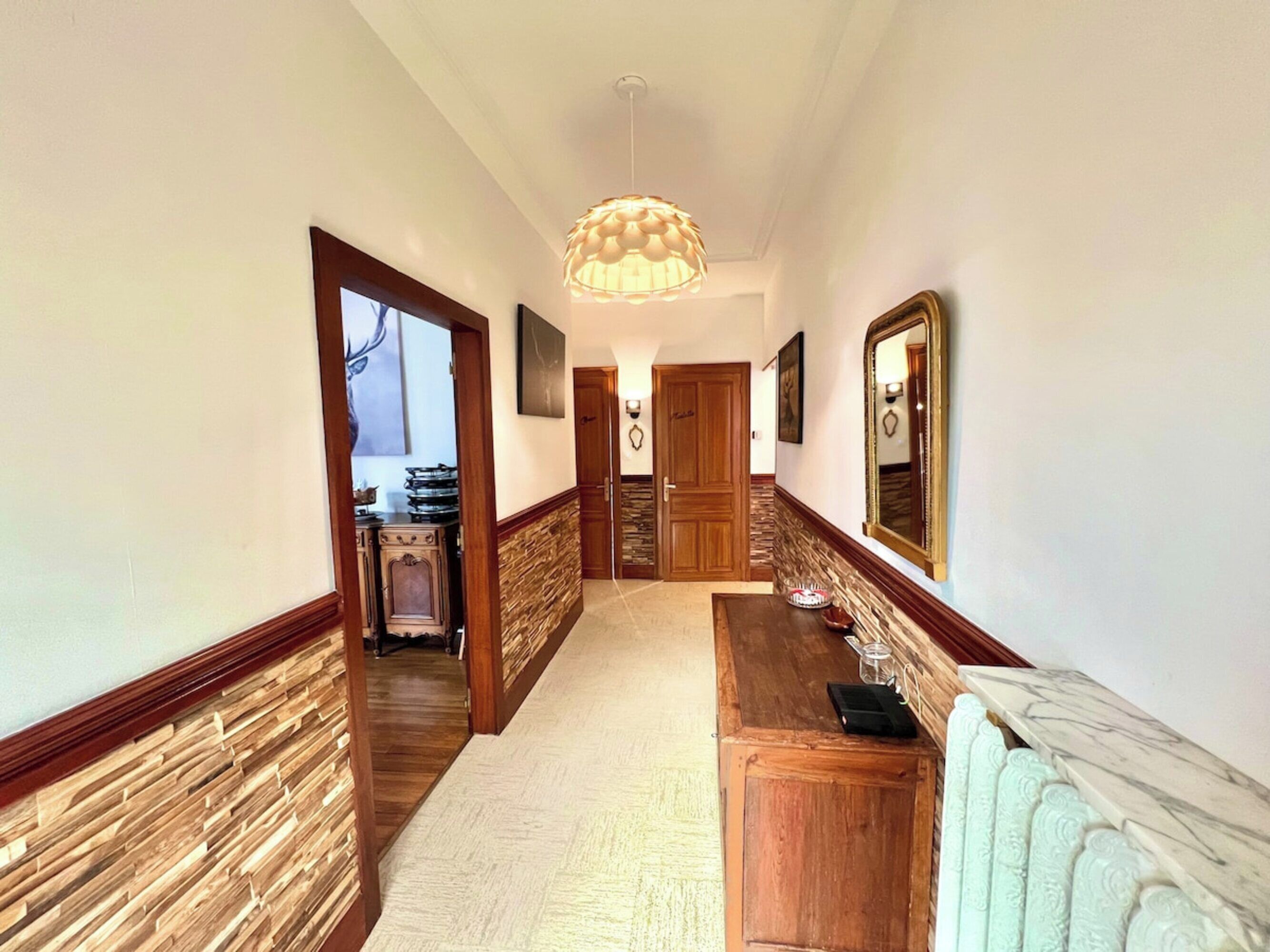 A hallway with a wooden floor, a marble bench, a wooden cabinet, and a large mirror.