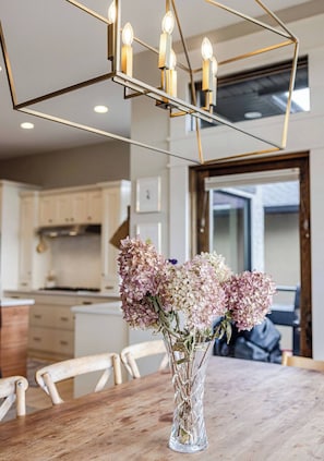 A dining table with a vase of flowers, a geometric light fixture, and a kitchen in the background.