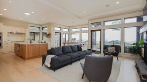 A spacious living area with a dark-colored sofa, a coffee table, and a view of the outdoors through large windows. - Bright open-concept home in Kelowna with lake views, high ceilings, fireplace. (Kelowna)