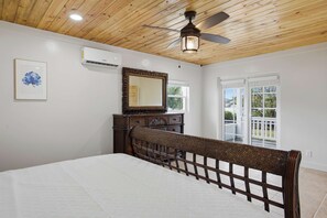 A bedroom with a wooden ceiling, a ceiling fan, a bed with a wooden headboard, a dresser, a mirror, and a window with a view.