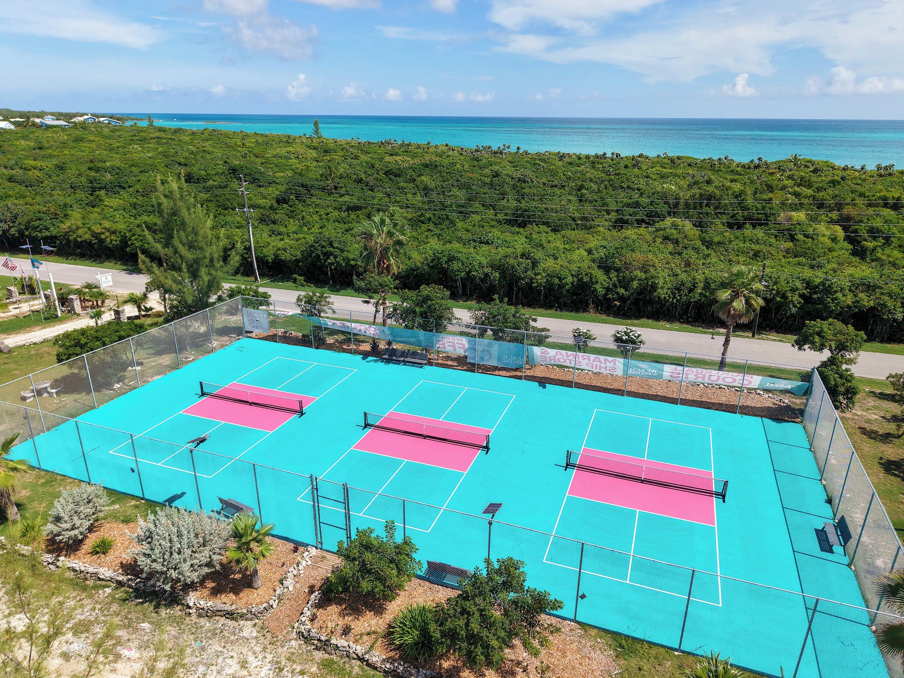 A tennis court with a net, surrounded by a fence, and a view of the ocean and greenery in the background.