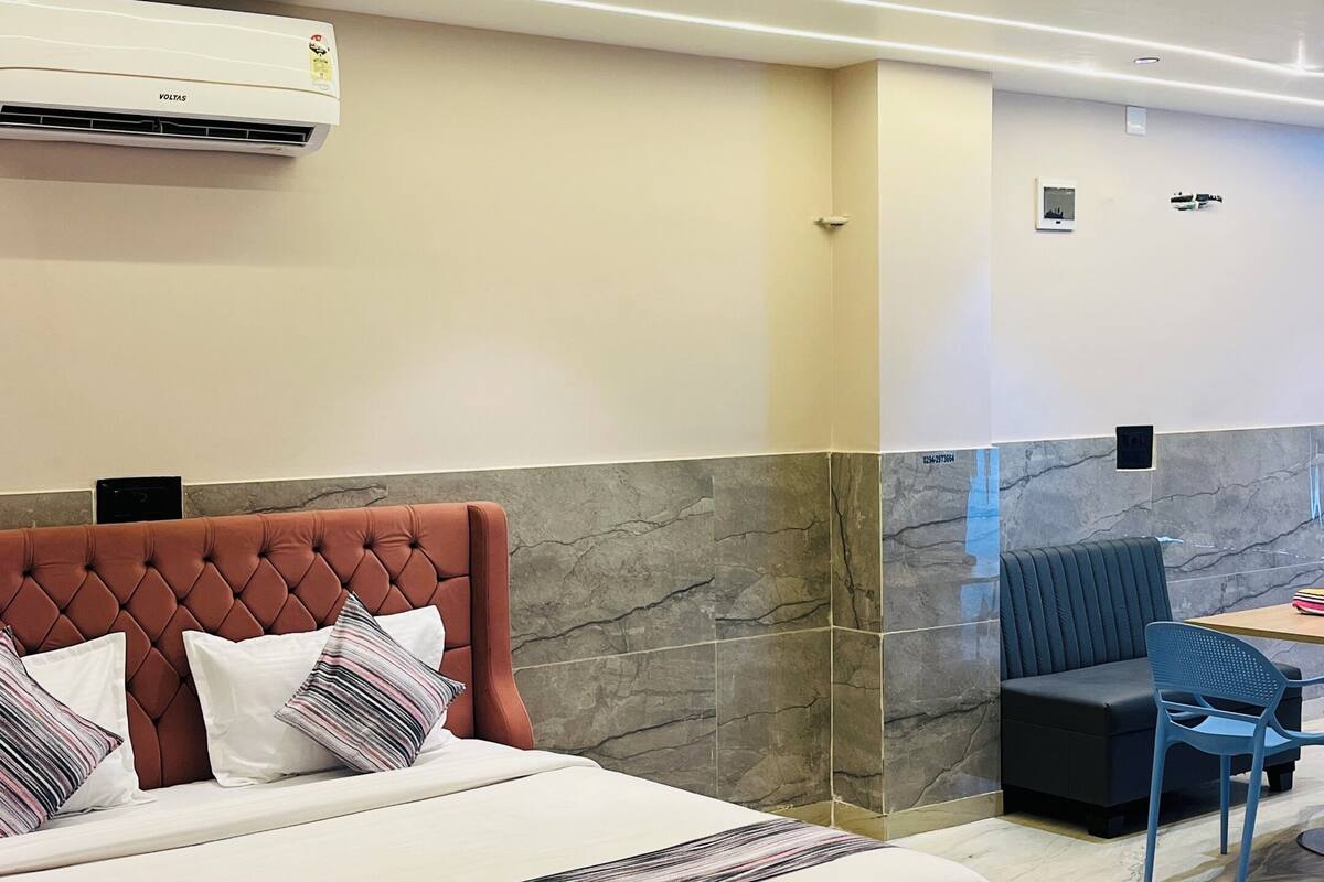 Una habitación de hotel moderna con una cama grande, ventilador de techo, equipo de aire acondicionado, zona de estar con sofá y sillones, y suelo de mármol.