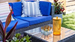 Un sofá azul con almohadones a rayas, una mesa de centro de vidrio con bebidas y una maceta de mimbre con flores rojas.