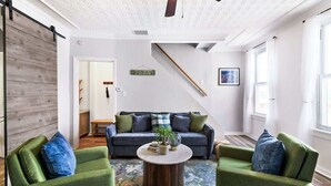 Una sala de estar con un sofá, dos sillones, una mesa de centro, techo de madera, una escalera y una puerta.