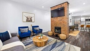 A living room with a fireplace, blue armchairs, a round coffee table, and a kitchen area in the background. - 2 King Beds w Hot Tub Near Butler St (Pittsburgh)