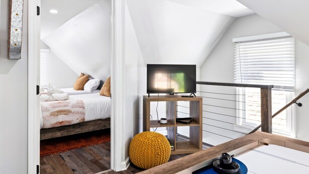A bedroom with a bed, a TV on a wooden stand, a yellow pouf, and a wooden staircase. - 2 King Beds w Hot Tub Near Butler St (Pittsburgh)