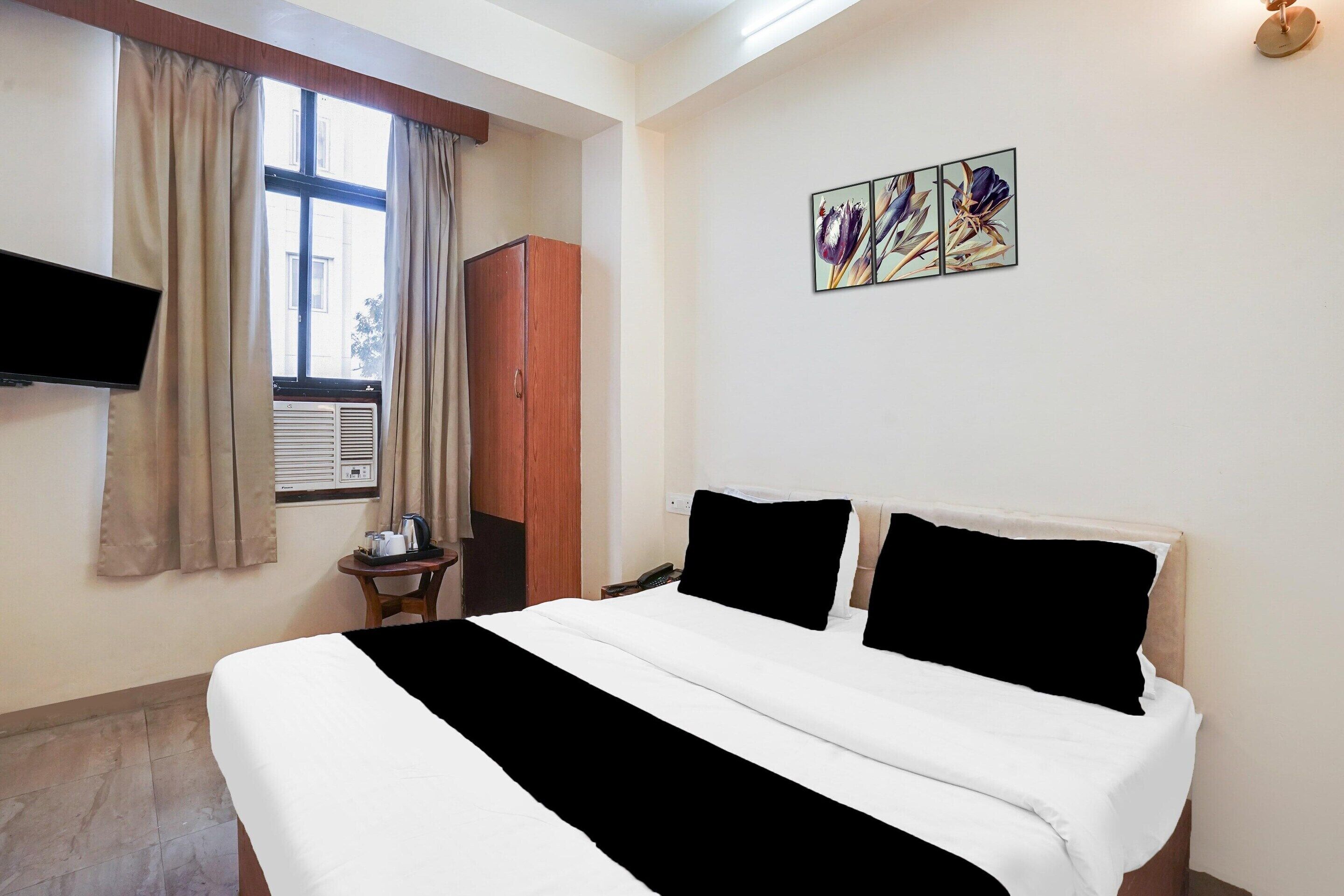 A hotel room with a bed, a television, a window with curtains, a wall-mounted artwork, and a wooden wardrobe.