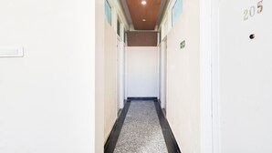 Ein schmaler Flur mit einem schwarz-weißen Musterboden, weißen Wänden und einer Decke mit eingelassener Beleuchtung. Eine Tür mit der Nummer 205 ist rechts zu sehen.