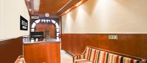 A hotel lobby with a reception desk, a clock, striped seating, and an exit sign.