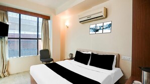Ein Hotelzimmer mit einem Bett, einem Sessel, einem Schreibtisch, einer Klimaanlage, einem an der Wand montierten Fernseher und einem Fenster mit Vorhängen.