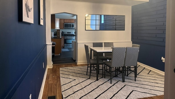 A dining area with a table and chairs, a kitchen in the background, and a modern light fixture.