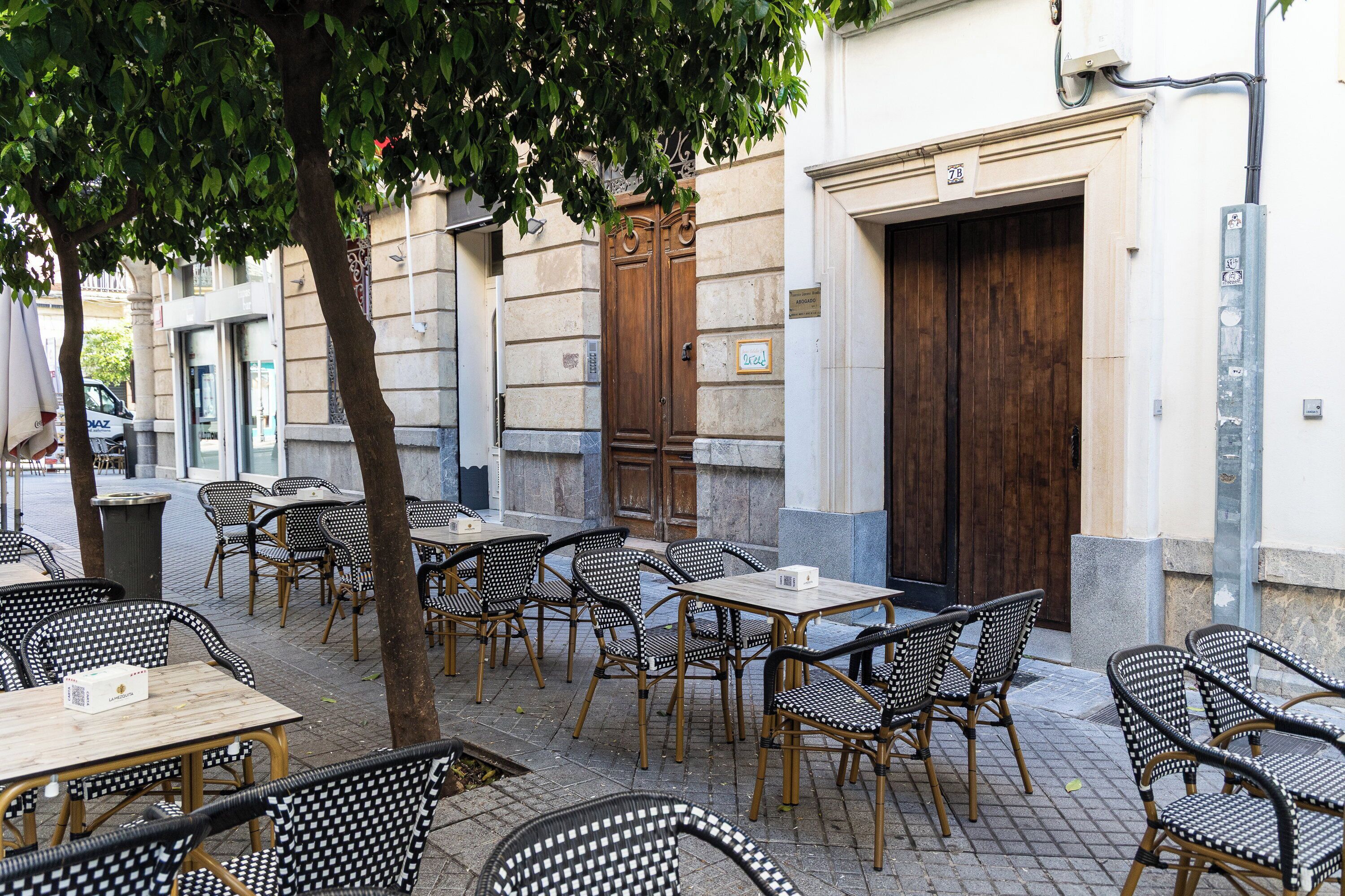 Un espace café en plein air avec des tables et des chaises, un arbre et un bâtiment doté d’une grande porte en bois et d’une enseigne.