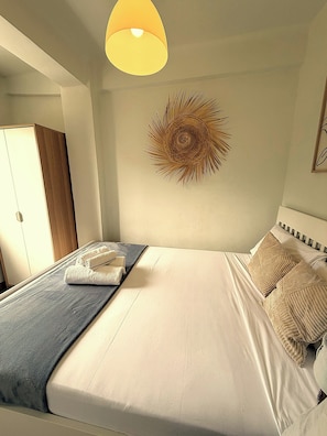 Cama bem arrumada com manta azul, um objeto decorativo na parede e um lustre.