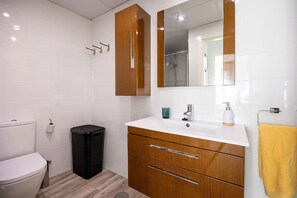 Ein Badezimmer mit weißen Fliesen, einem hölzernen Waschtisch mit Spiegel, einer Toilette und einem Mülleimer.