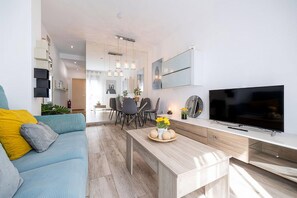 Ein modernes Wohnzimmer mit einer Couch, einem Couchtisch mit Blumen, einem Fernseher und einem Essbereich mit Stühlen und Tisch.