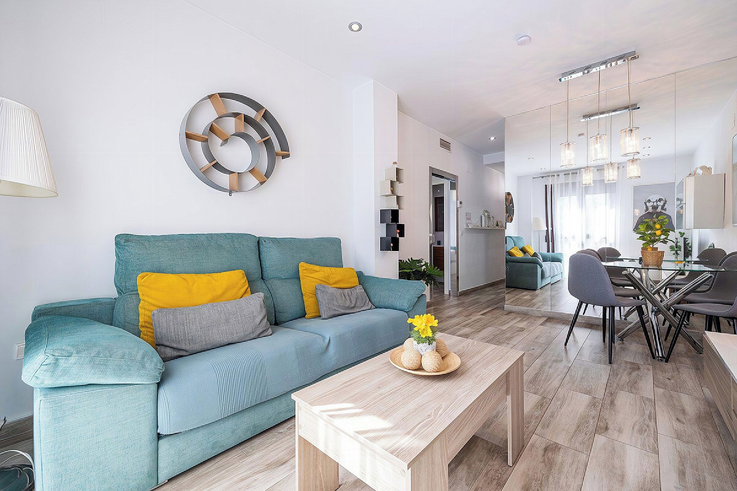 A modern living room with a teal sofa, a wooden coffee table, and a dining area with a glass table and chairs.