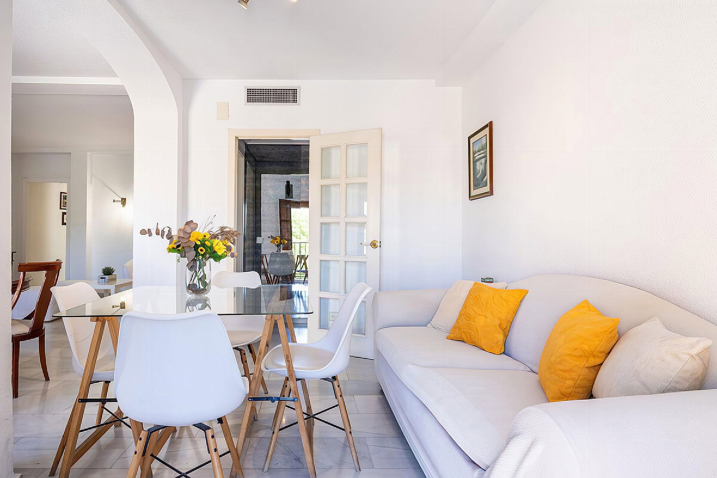 A modern living room with a glass dining table, white chairs, and a sofa with yellow cushions.