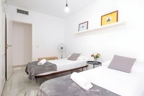 Ein Hotelzimmer mit zwei Betten, einem Nachttisch mit Lampe, einem Wandventilator und einem Regal mit gerahmten Bildern.