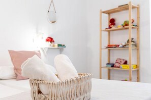 Una habitación con una cama, una cesta de mimbre con toallas, una estantería de madera y un espejo colgado.