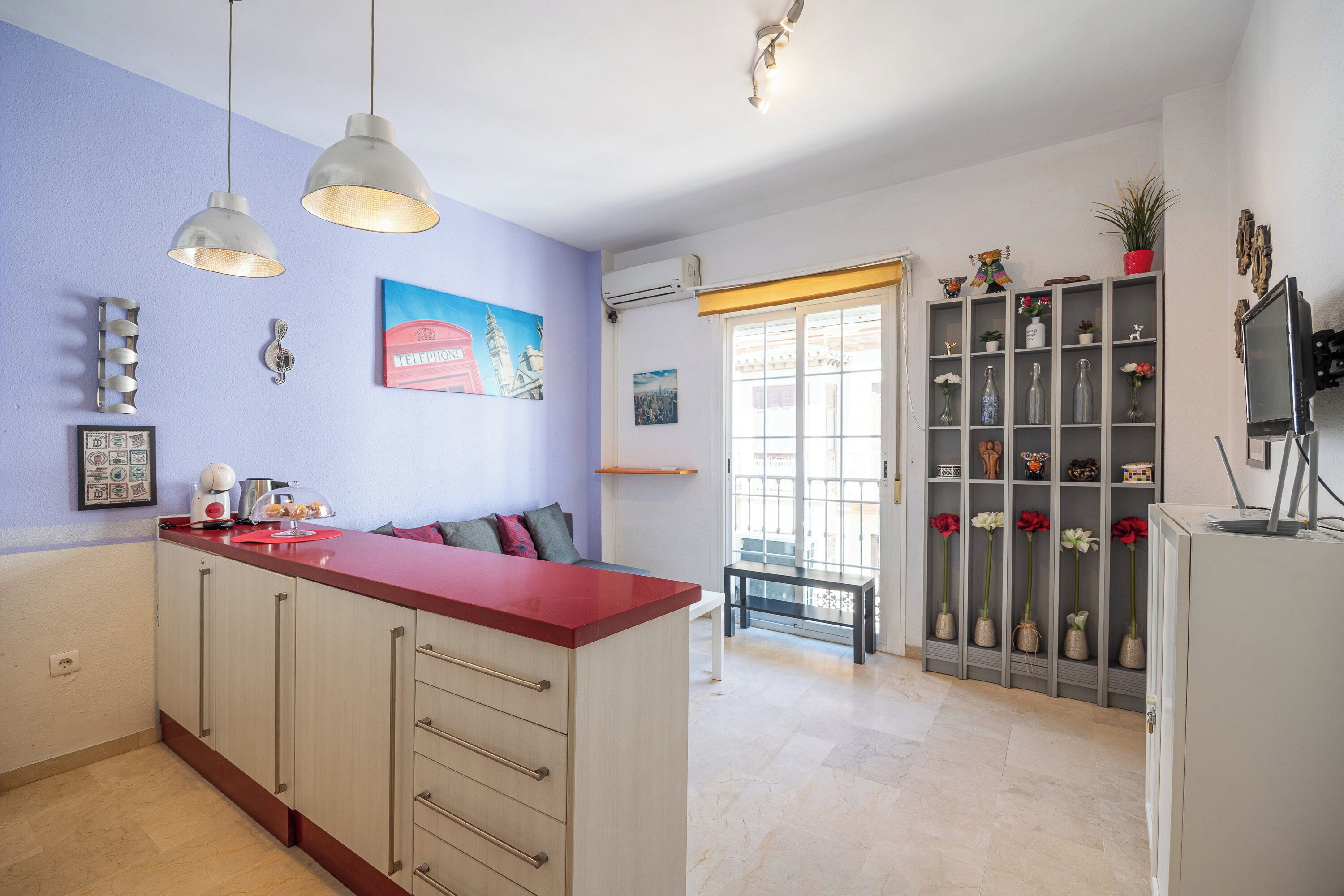 A kitchen with a red countertop, a dining area with a bench, and a shelving unit with decorative items.