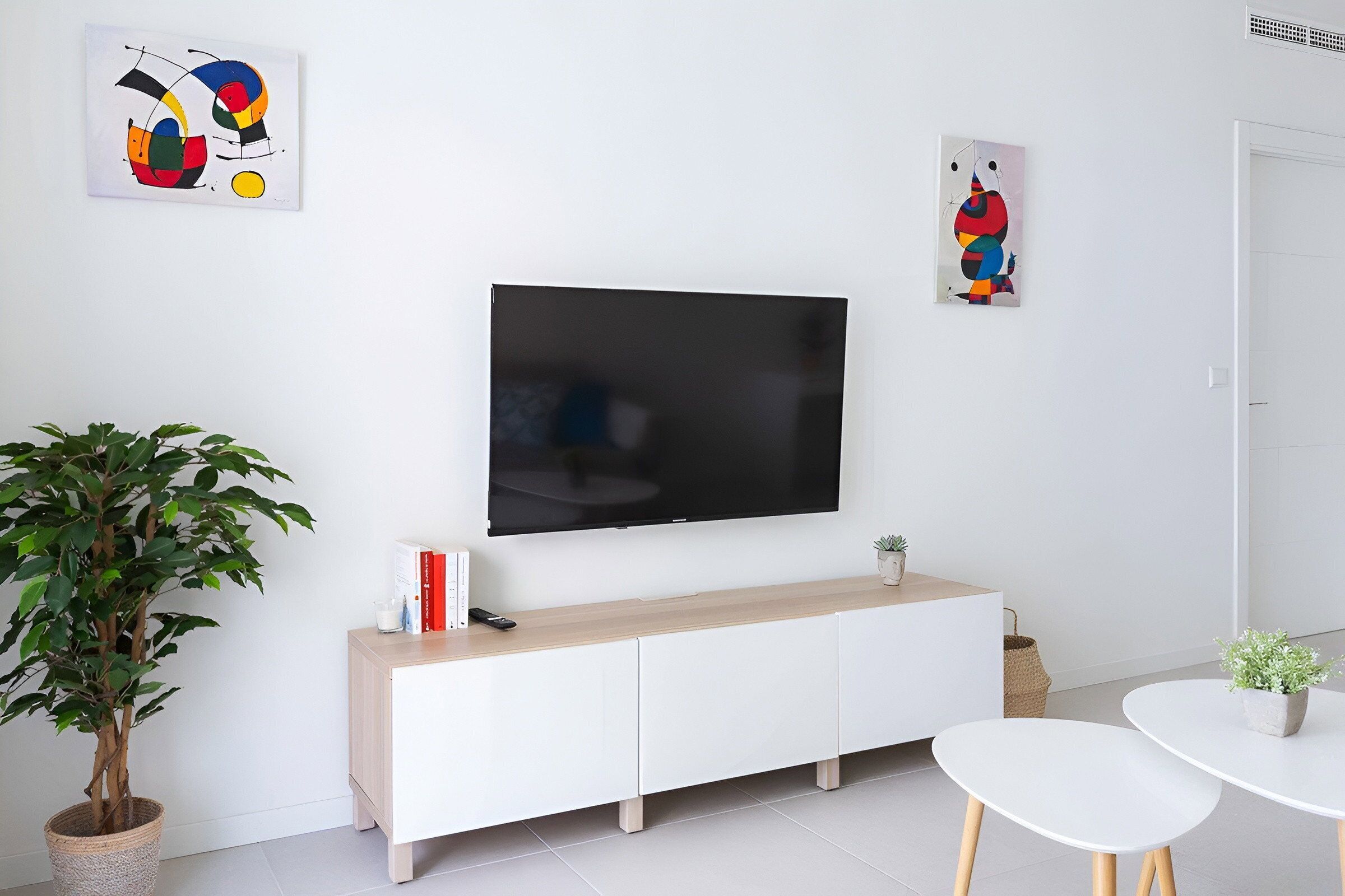 A modern living room with a flat-screen TV mounted on the wall, a white cabinet, a potted plant, and two white tables.