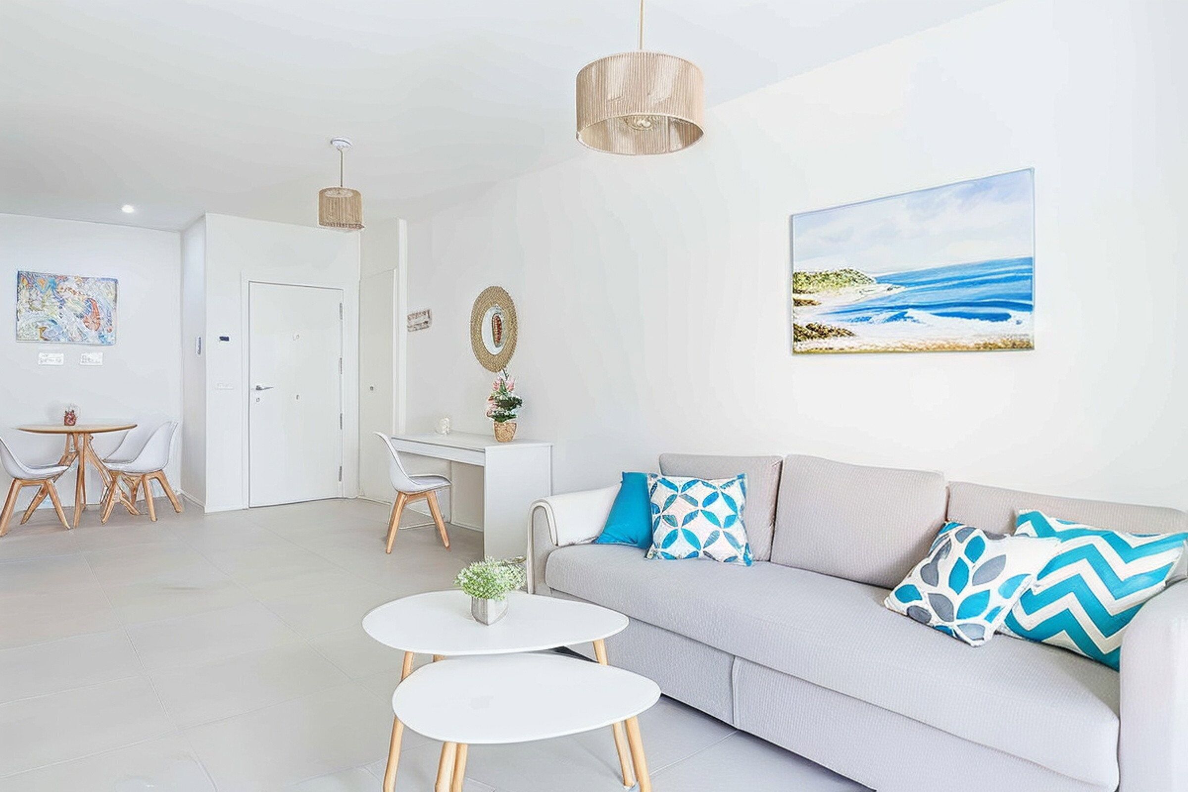Ein modernes Wohnzimmer mit einem grauen Sofa, zwei weißen Beistelltischen, einem Essbereich mit weißem Tisch und Stühlen, einem Schreibtisch und einem Gemälde einer Strandansicht.