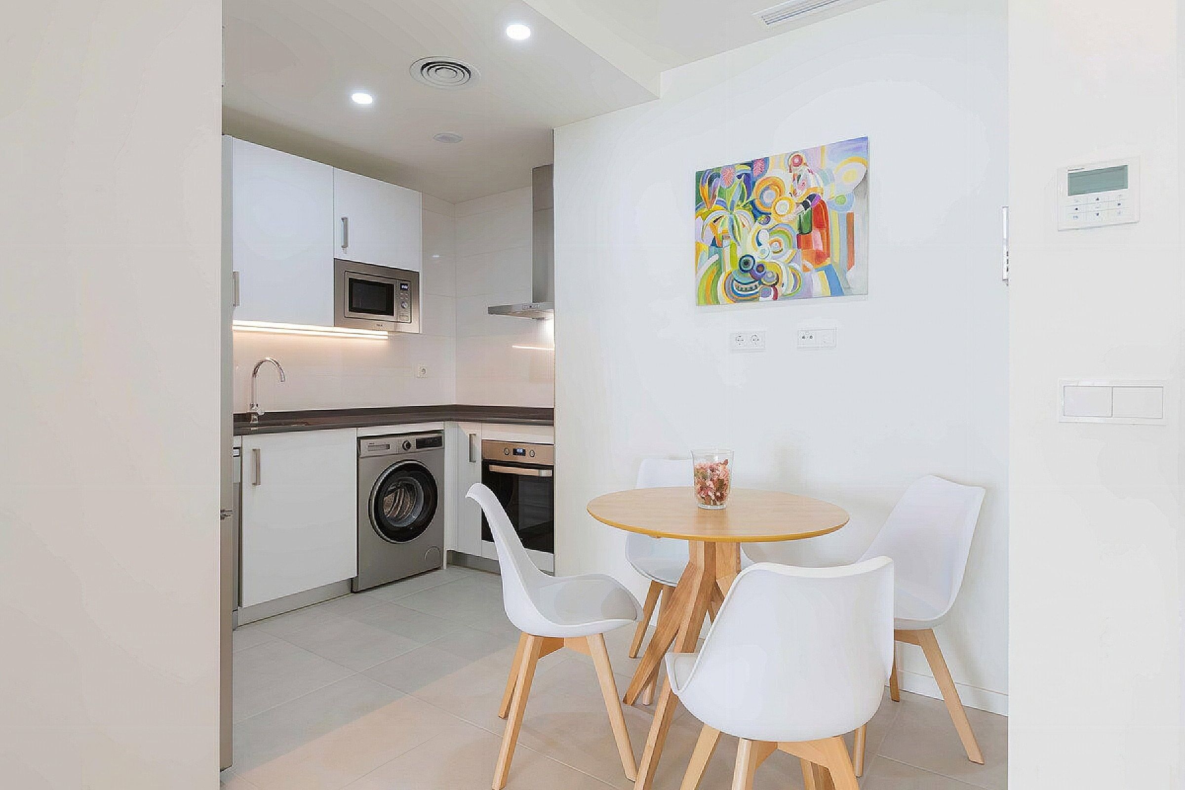 A modern kitchen with built-in appliances, a dining area with a round table and white chairs, and a colorful abstract painting on the wall.