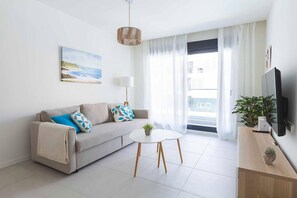 Ein modernes Wohnzimmer mit einer Couch, einem kleinen runden Tisch, einer Lampe, einem Fernseher und einem Fenster mit Vorhängen.