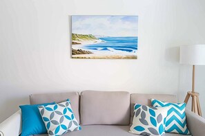 Ein Wohnzimmer mit einem beigen Sofa, zwei gemusterten Kissen, einem Gemälde mit Strandmotiv und einer Stehlampe.