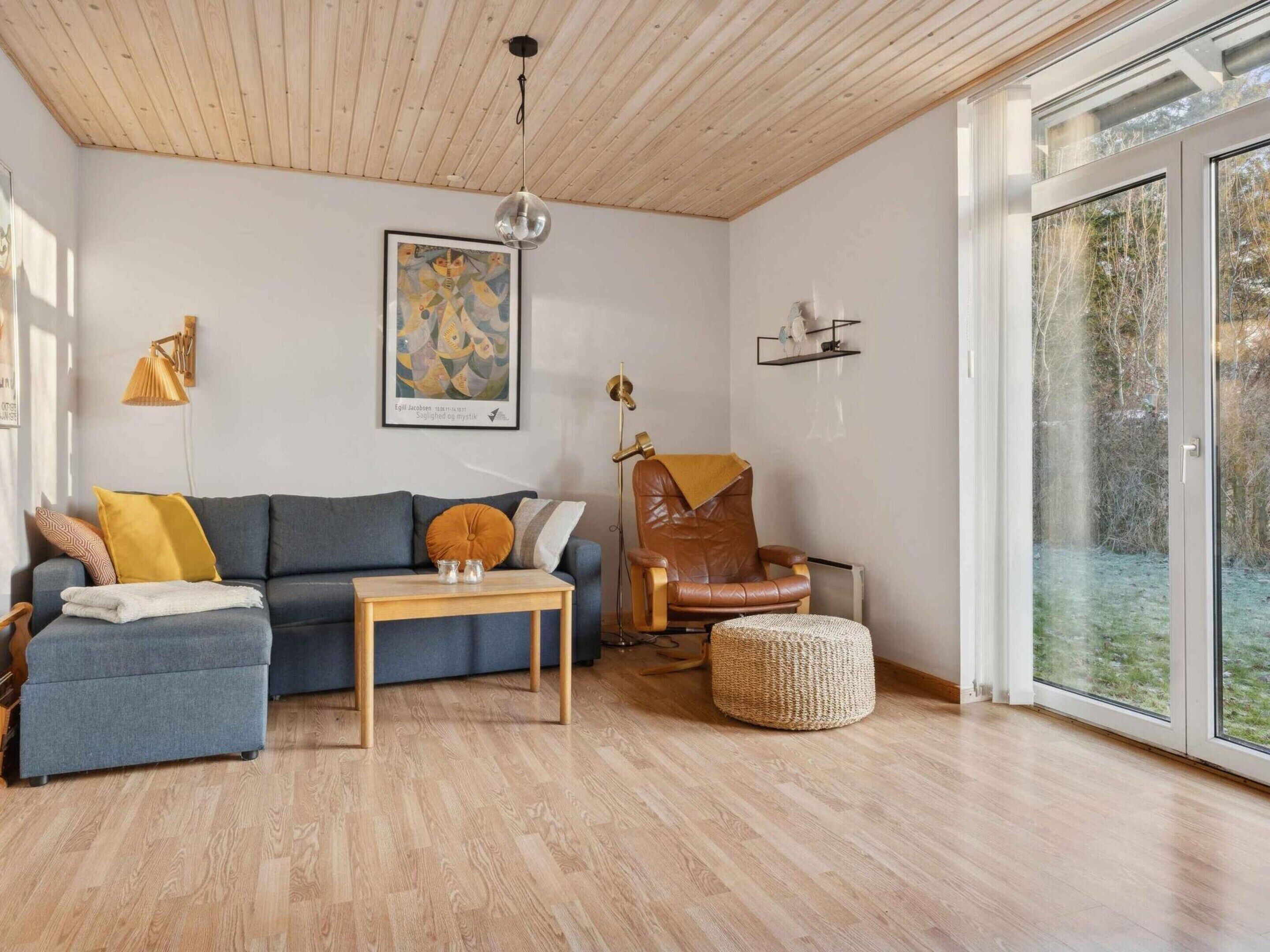 A living room with a wooden ceiling, a grey sofa, a brown leather chair, a small table, and a large window.