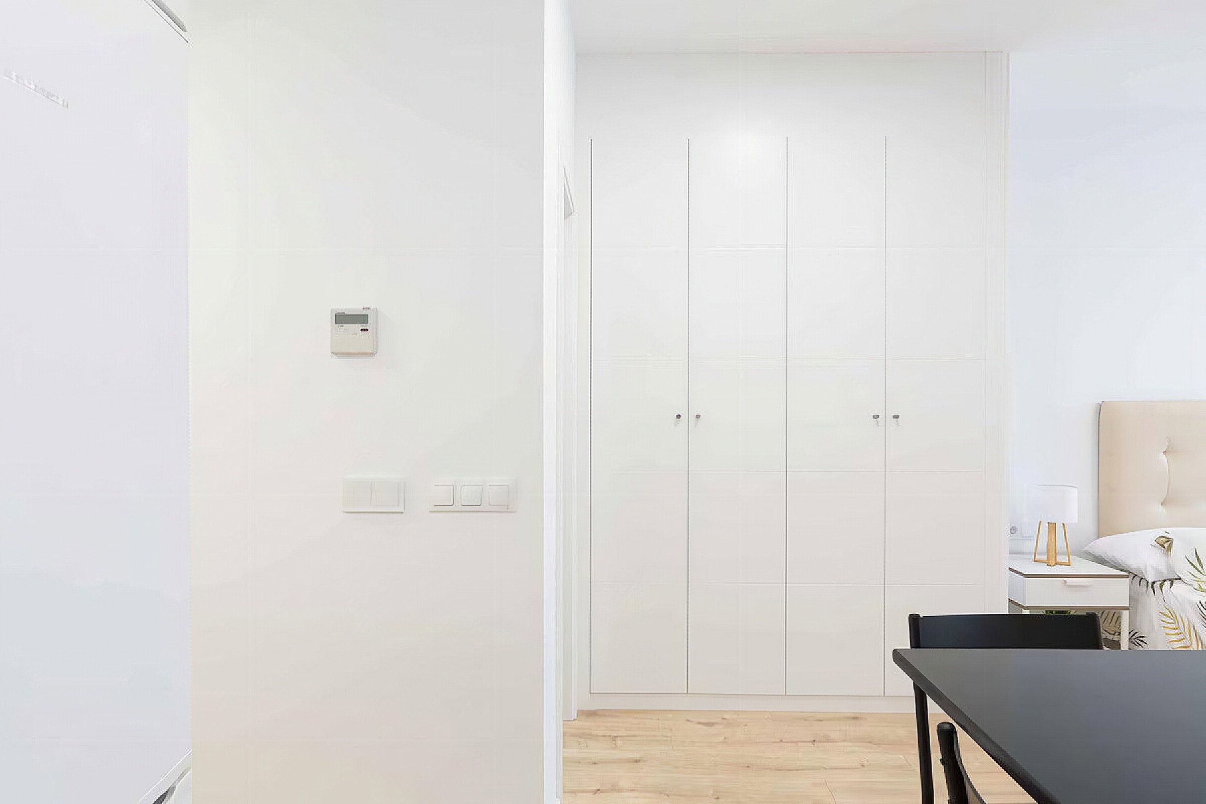 A minimalist room with a white door, a small bedside table with a lamp, and a wooden floor.