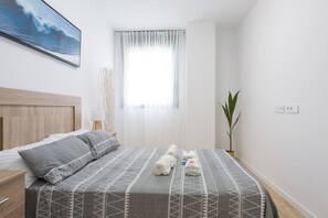 A neatly made bed with a patterned bedspread, a bedside table with a lamp, a potted plant, and a painting on the wall. - Cosmopolitan Apt & Malaga Beach (Málaga)