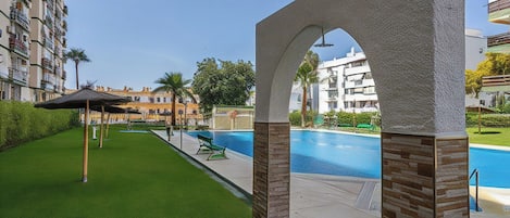 A swimming pool area with a clear sky, a well-maintained lawn, and a building with balconies in the background.