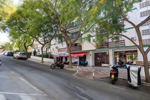 Een straat met geparkeerde auto's en motoren, bomen langs de stoep en gebouwen met winkelruiten.