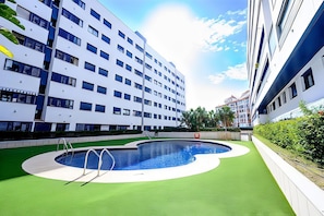 A modern building with multiple floors, a swimming pool with a green deck, and a clear sky. - Tradiciones Marineras, Puerto y Plaza de Toros (Estepona)