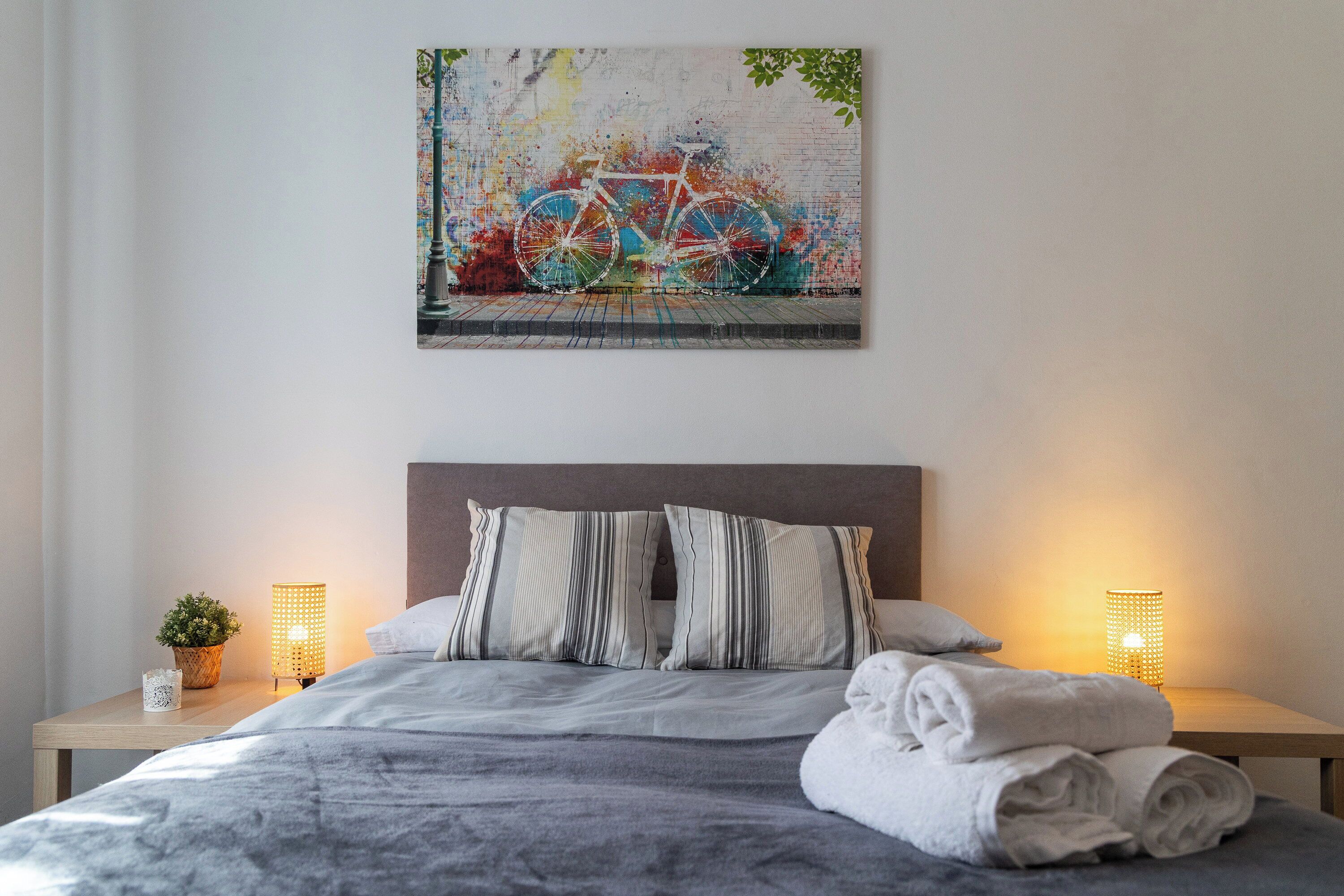 Ein ordentlich bezogenes Bett mit gestreiften Kissen, ein gefaltetes Handtuch und ein an der Wand hängendes Bild von einem Fahrrad.