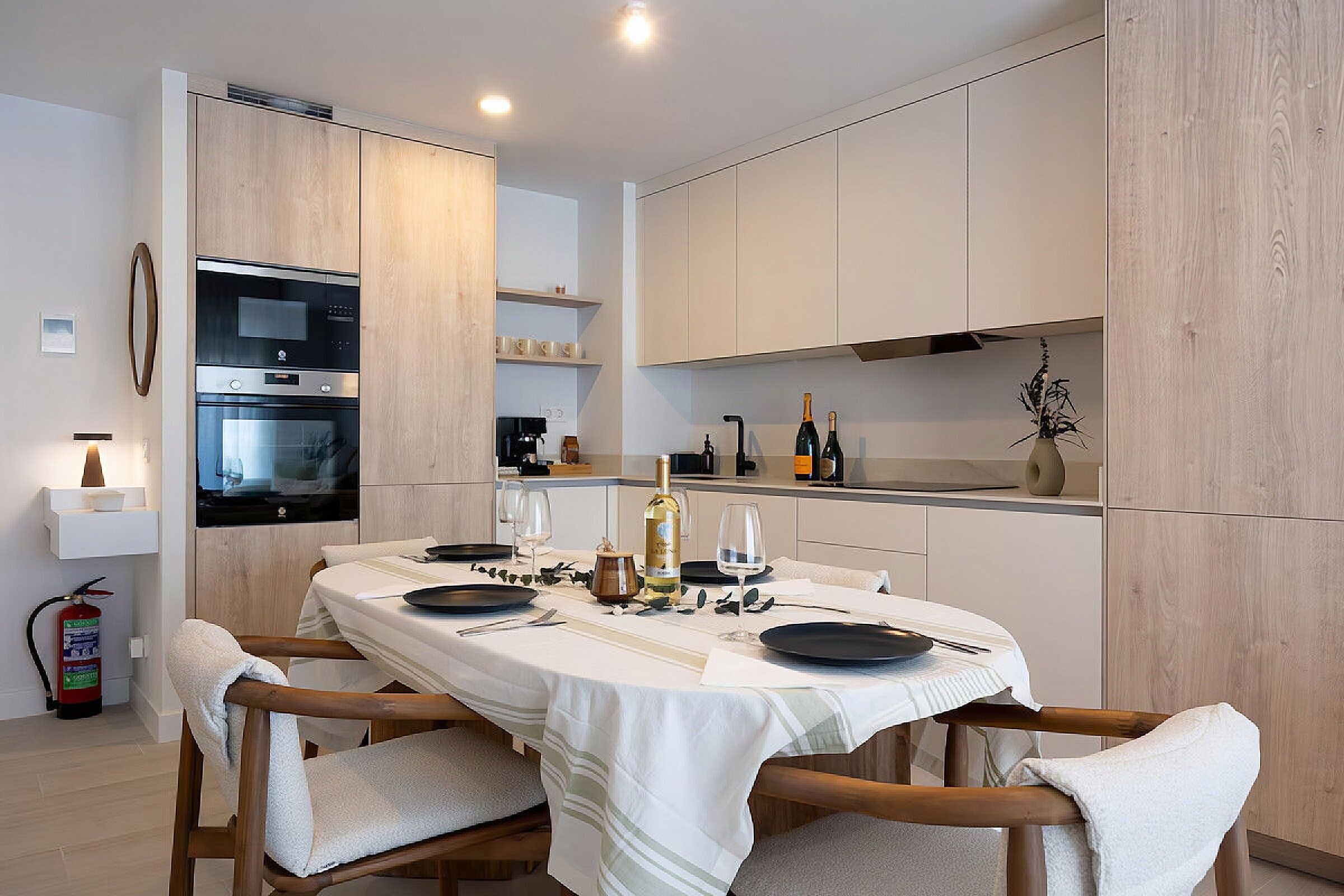 A modern kitchen with a dining table set for two, equipped with a coffee machine, wine bottles, and a decorative vase.