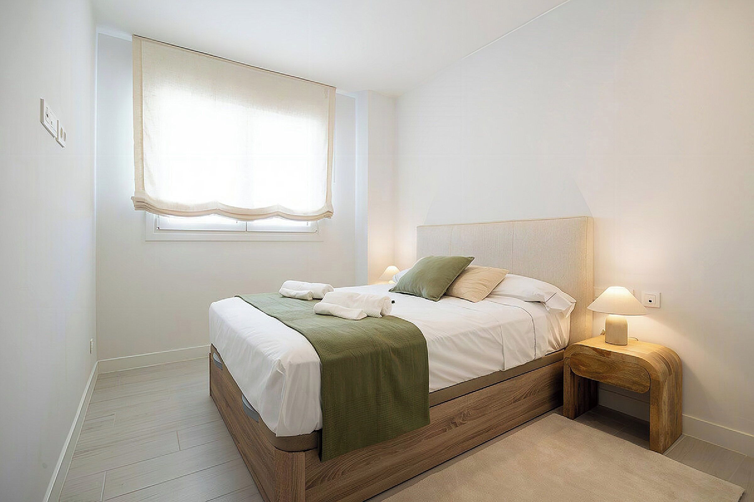 A bedroom with a bed, bedside lamps, a window with curtains, and a wooden floor.