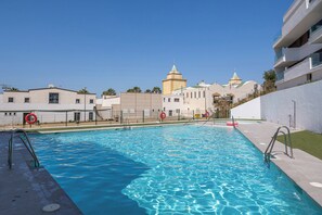 En swimmingpool med klart, blåt vand, omgivet af et hegn, ved siden af en bygning med en hvid facade og altaner.