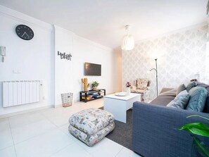 Ein modernes Wohnzimmer mit einer weißen Heizung, einer Uhr, einem Flachbildfernseher, einem Kaktus, einer kaktusförmigen Skulptur, einem gemusterten Hocker, einem blauen Sofa und einem gemusterten Sessel.