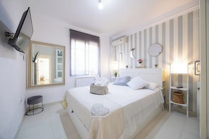 A bedroom with a bed, a television mounted on the wall, a window with curtains, a radiator, and a bedside table with a lamp. - Cordoba in Bloom Steps from Alcazar & Mosque (Cordoba)