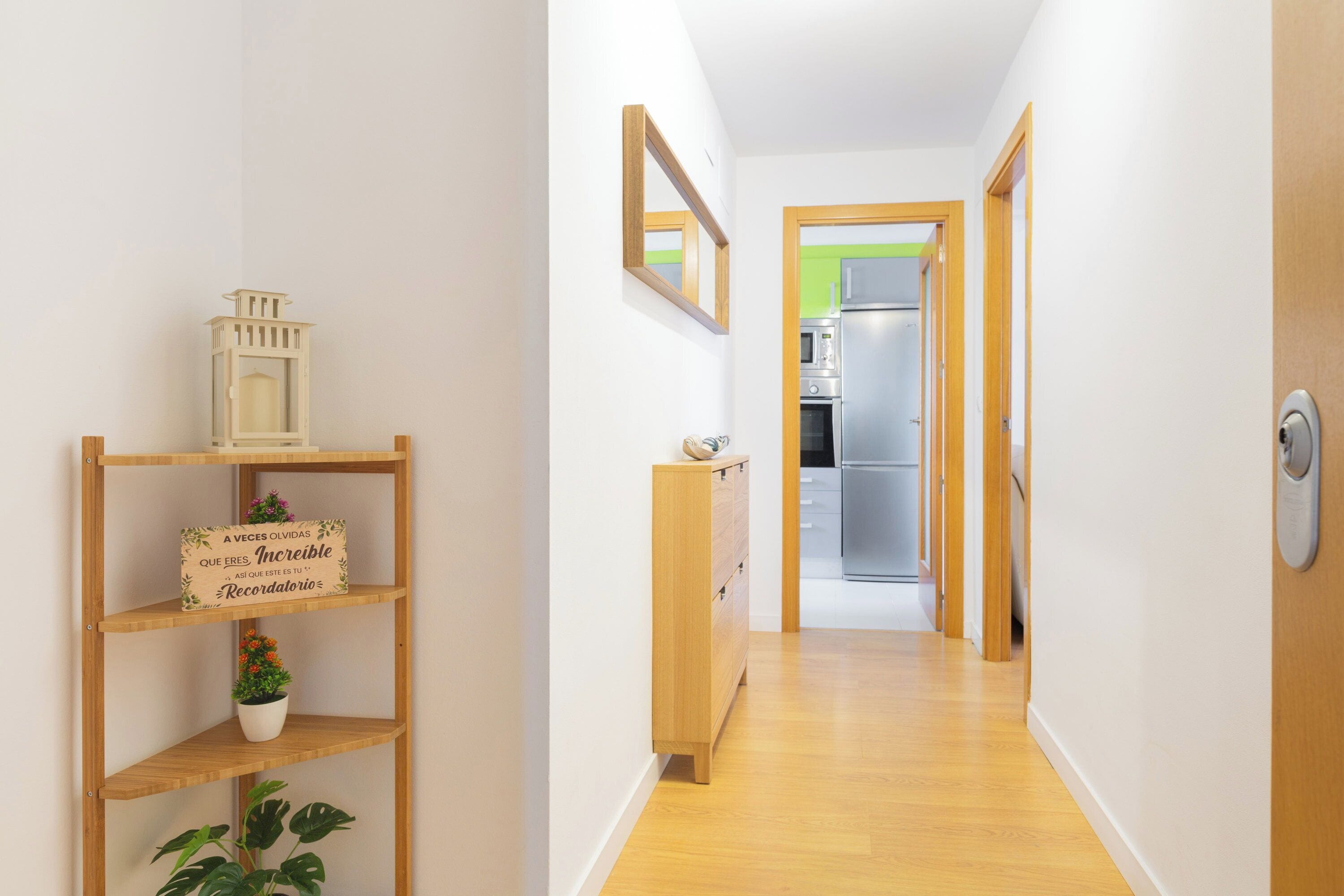 A hallway with wooden flooring, a wooden shelf with decorative items, and a kitchen visible through an open door.