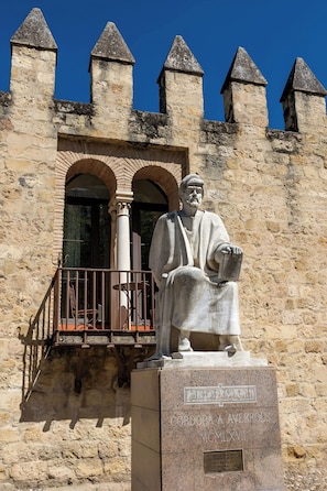 Ein Steinhaus mit einer Statue einer sitzenden Person, einem Balkon und blauem Himmel.