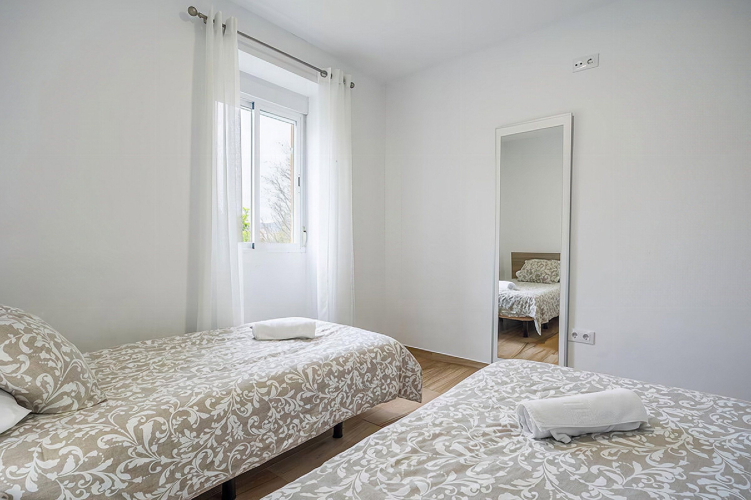 A bedroom with two beds, a window with curtains, a mirror, and a door.