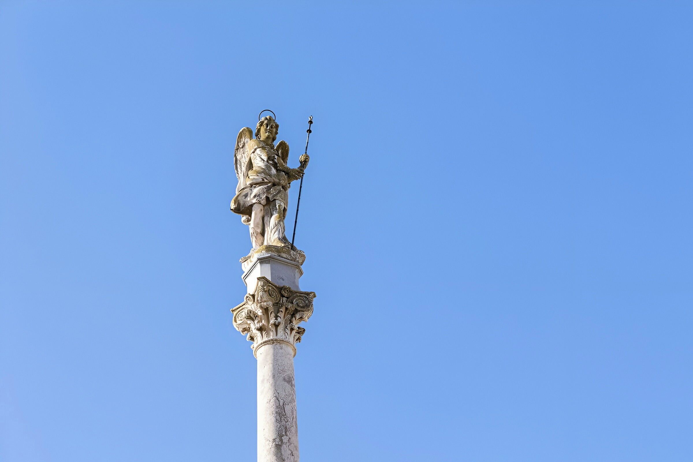 Una estatua en la cima de una columna con tallas elaboradas, al pie de un cielo azul despejado.