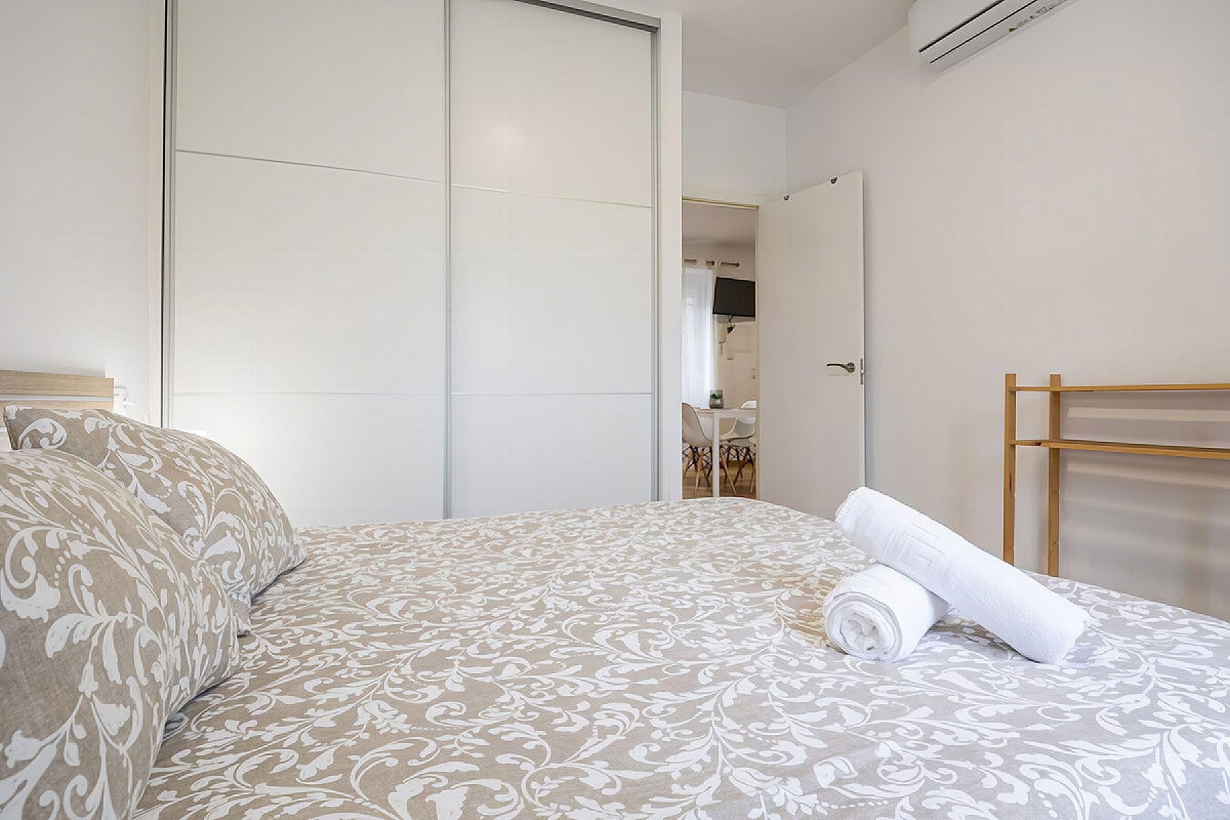 A hotel room with a bed, a wooden side table, a mirror, and a view of a dining area.