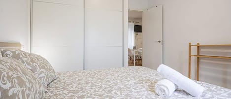 A hotel room with a bed, a wooden side table, a mirror, and a view of a dining area.