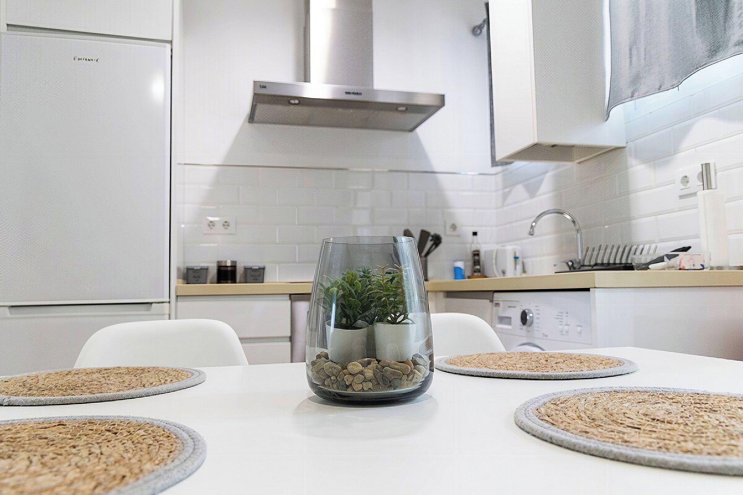 Una cocina moderna con electrodomésticos blancos, una mesa de vidrio con un centro de mesa de planta y un diseño limpio y minimalista.