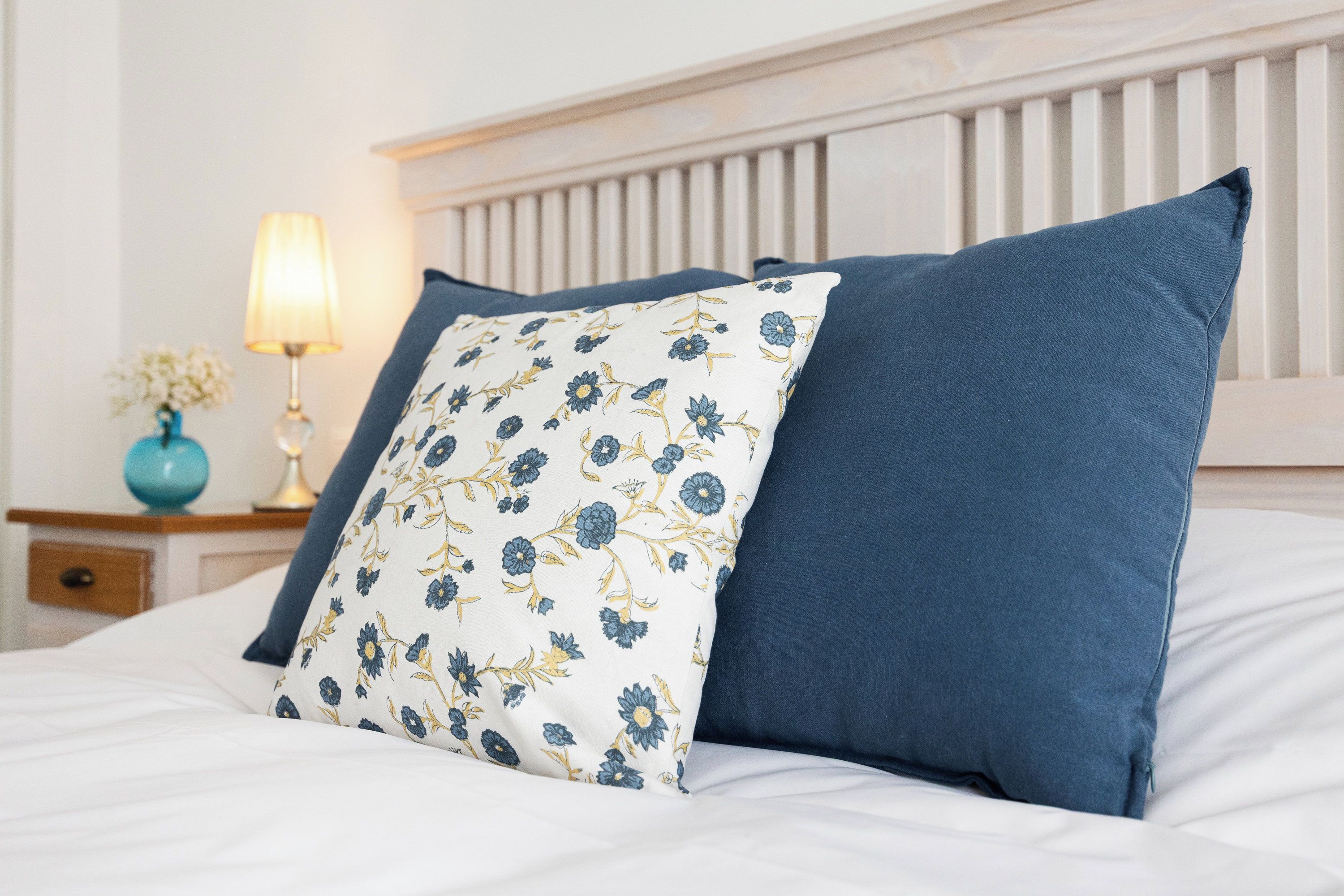 Una cama con dos almohadas, una con estampado floral y otra azul lisa, frente a un cabecero blanco.